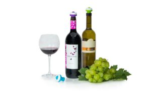 Steklenici vina z vakuumskimi zamaški, natočen kozarec črnine in grozdje na belem ozadju.